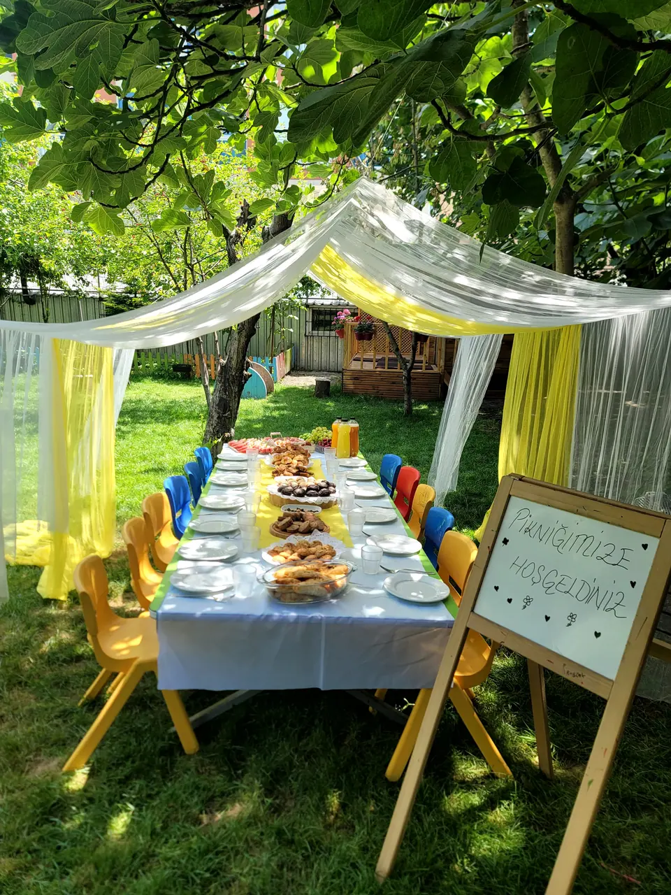 Ağaçların altında sarı ve beyaz tüllerle süslenmiş piknik sofrası